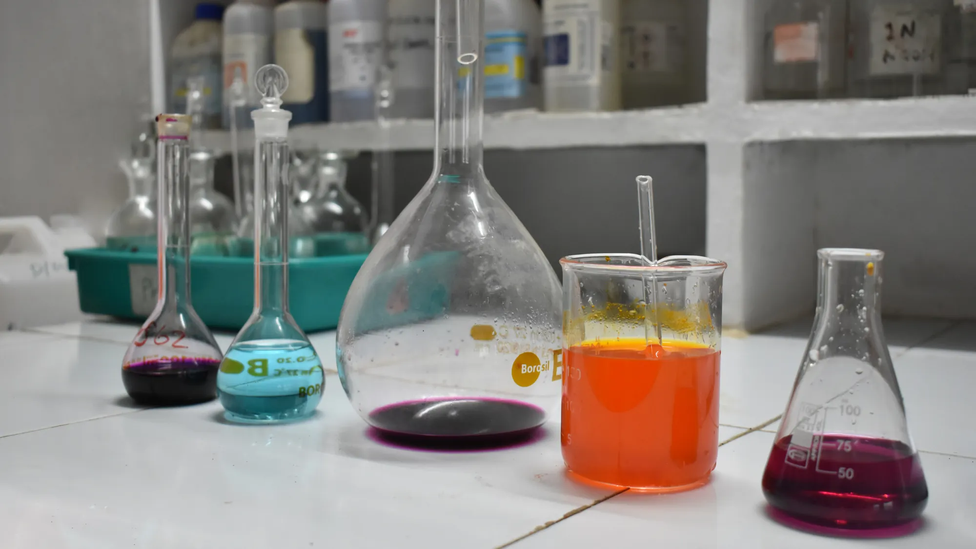Laboratory glassware holding coloured analytical reagents on a laboratory bench, with chemical reagent bottles on shelves behind